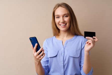 Happy young woman holding smartphone and credit card against beige background