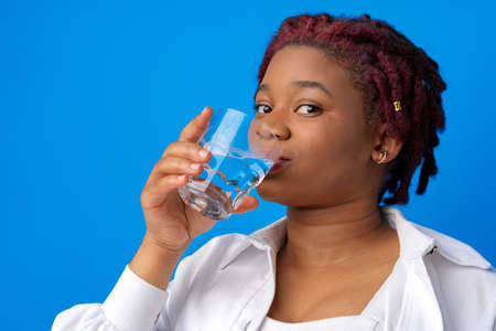 Young african woman drinking water from a glass against blue backgroundの写真素材