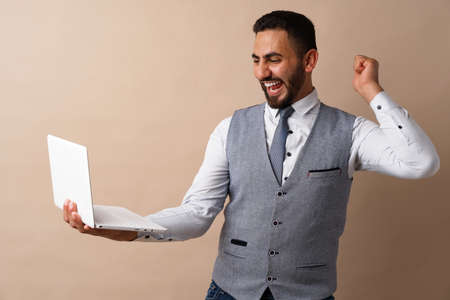 Cheerful young handsome Arab man working on laptop while standing against beige background