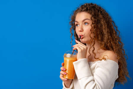 Beautiful curly-haired woman drinking orange juice against blue background