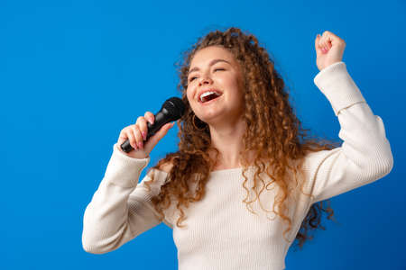 Young joyful curly-haired woman singing in microphone against blue background