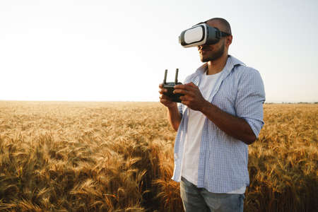 Young man standing in a wheat field at sunset in virtual reality glasses