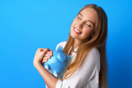 Beautiful smiling young woman with piggy bank on blue backgroundの写真素材