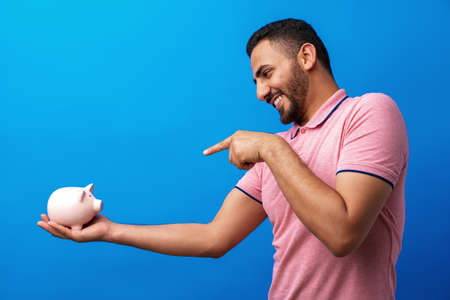 Handsome very excited hispanic man holding piggy bank against blue background