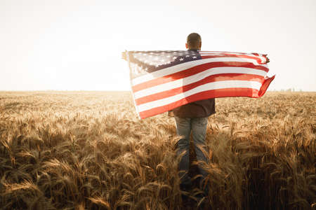 Young man holding American flag on back while standing in wheat fieldの写真素材