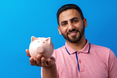 Handsome very excited hispanic man holding piggy bank against blue backgroundの写真素材