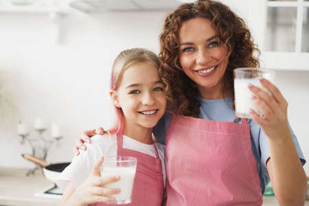 Mother and daughter smiling and holding glasses of milk in kitchenの写真素材