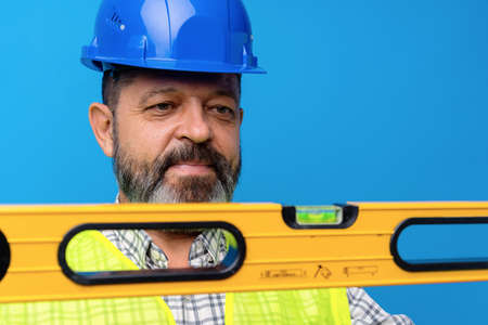 Portrait of male handyman in blue hardhat holding building level against blue background
