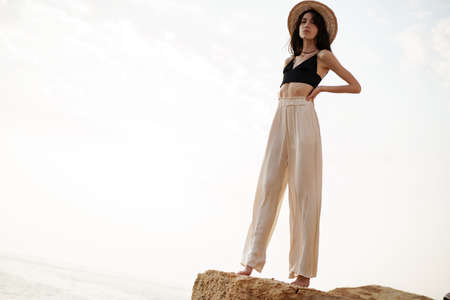 Young stylish woman standing on cliff at the sea at sunset