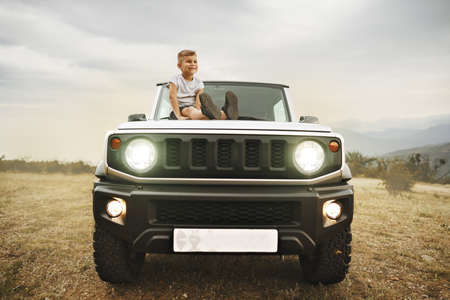 Little boy sitting on a hood of an off-road car parked in mountains