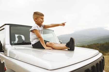 Little boy sitting on a hood of an off-road car parked in mountainsの写真素材
