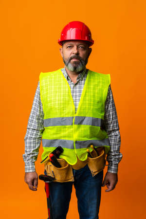 Middle-aged bearded man builder in uniform and hardhat with tools against orange backgroundの写真素材