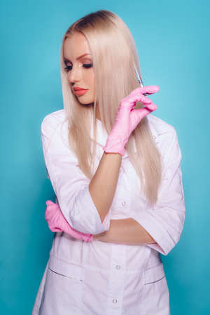 Beautiful young woman doctor beautician in medical uniform holding syringe on blue background. Beauty and cosmetic injection conceptの写真素材