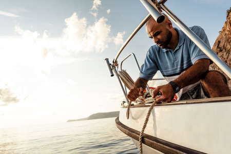 Young african american sailor tying ropes on sailboat in the sea on sunsetの写真素材