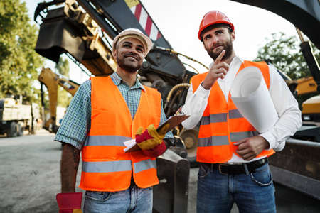 Two men engineers discussing their work standing against construction machinesの写真素材