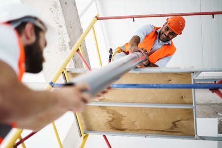 Two men builder in uniform drag the pipe on scaffolding at construction siteの写真素材