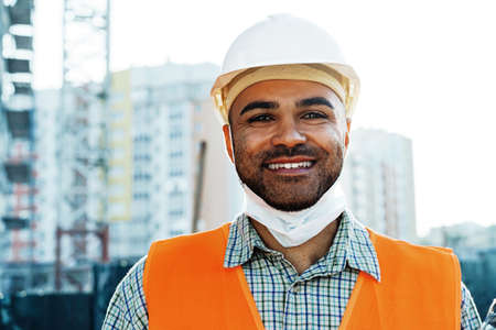 Portrait of mixed race man builder in workwear and hardhat wearing medical mask, close upの写真素材