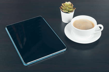 Digital tablet with black screen, coffee mug and small plant on a working table