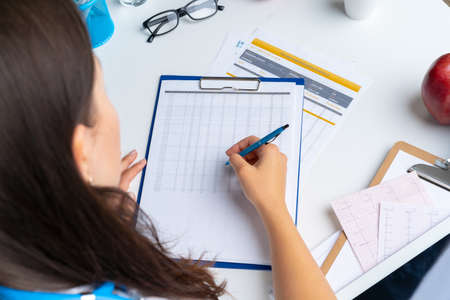 Woman doctor taking notes on clipboard while sitting at her table in officeの写真素材