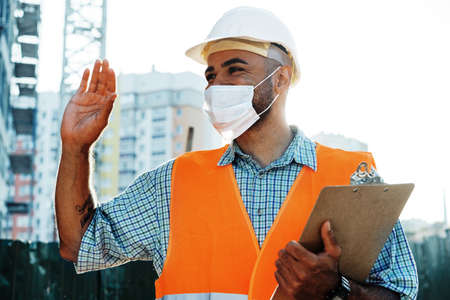 Portrait of mixed race man builder in workwear and hardhat wearing medical mask, close upの写真素材