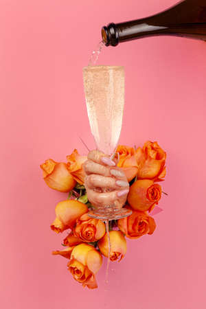 Female hand with manicure pouring champagne into a glass through a hole in a torn paper decorated with rosesの写真素材