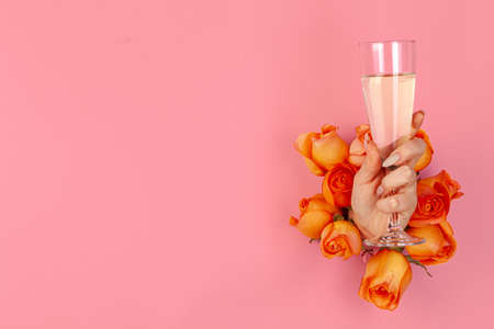 Woman holds a glass of champagne in her hand put through a hole in pink paper with fresh rosesの写真素材