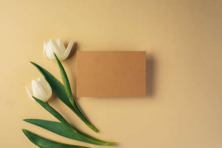 Craft paper card surrounded by fresh tulips on beige background