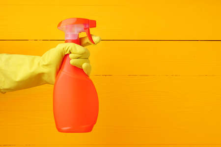 Hand in glove holding household detergent spray against yellow wooden backgroundの写真素材