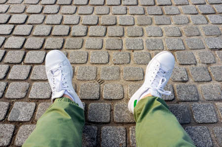 Male legs in green trousers and white sneakers on cobblestone pavement road