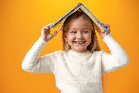 Little girl holding book on her head and looking at camera over yellow background