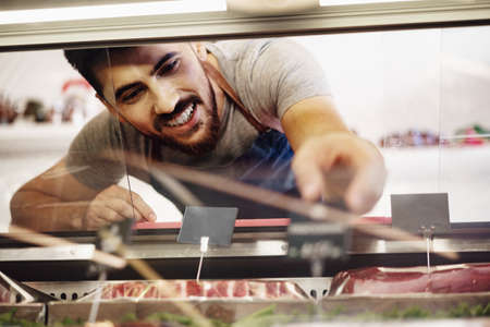 Young man butcher arranging meat products in display case of butcher shopの写真素材