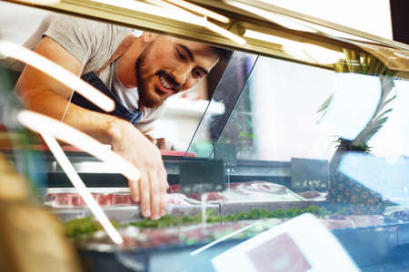 Young man butcher arranging meat products in display case of butcher shopの写真素材