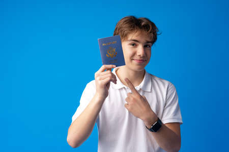 Portrait of glad handsome boy showing his passport going on vacation against blue backgroundの写真素材