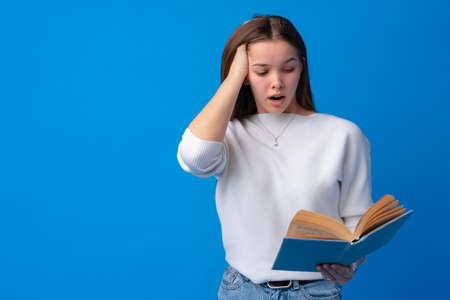 Young beautiful woman holding book scared and amazed on blue background in studioの写真素材