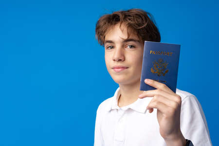 Portrait of glad handsome boy showing his passport going on vacation against blue backgroundの写真素材