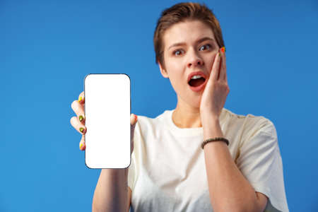 Portrait of a young woman showing blank screen mobile phone while standing over blue backgroundの写真素材