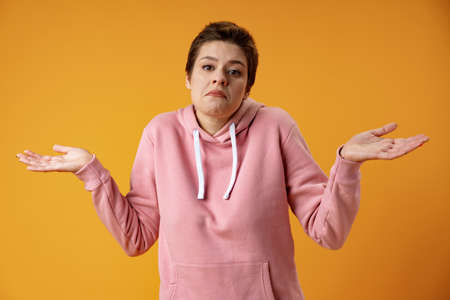 Studio portrait of awkward young woman feeling confused and puzzled shrugs shoulders over yellow...