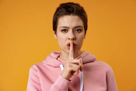 Serious young pretty woman showing silence gesture over yellow backgroundの写真素材