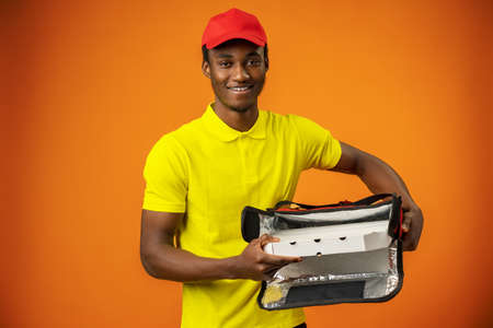Smiling black male courier wearing uniform holding box in orange studio