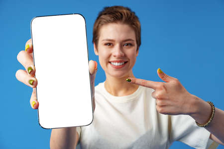 Portrait of a young woman showing blank screen mobile phone while standing over blue backgroundの写真素材