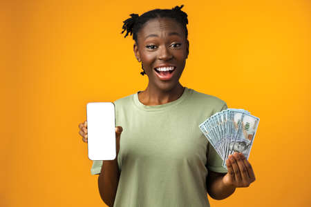 Young black woman holding white screen smartphone and money in yellow studioの写真素材