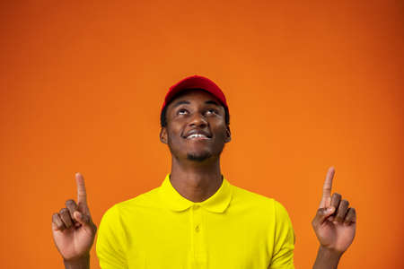 Delivery man in yellow t-shirt and red cap smiling and pointing finger up against orange background