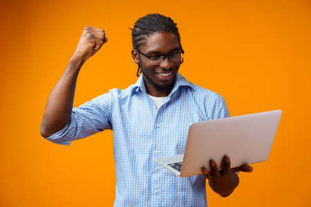 Young smiling african man standing and using laptop computer over yellow backgroundの写真素材