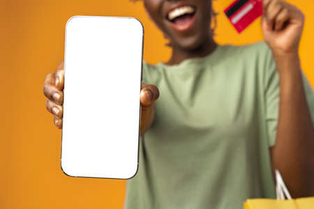 Smiling young afro girl holds smart phone and credit card in yellow studio