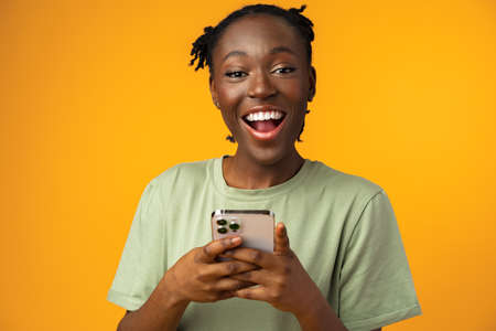 Happy smiling african american girl using her smartphone in yellow studio.
