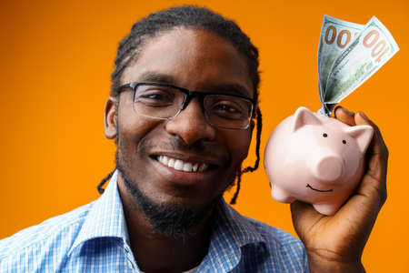 Young black african american man holding a piggy bank against orange background.の写真素材