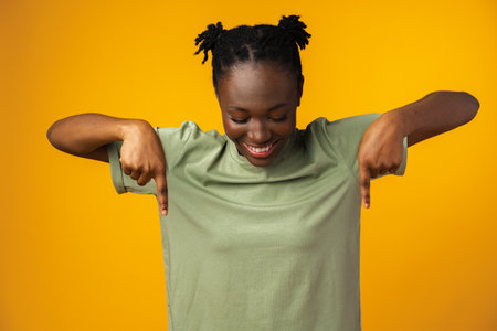 Cheerful Afro woman points to copy space in yellow studioの写真素材