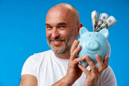 Middle-aged man holding piggy bank against blue backgroundの写真素材