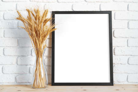 Vase with ears of wheat and photo frames against brick wall
