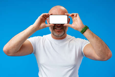 Middle-aged man holding blank smartphone on his eyes in studio